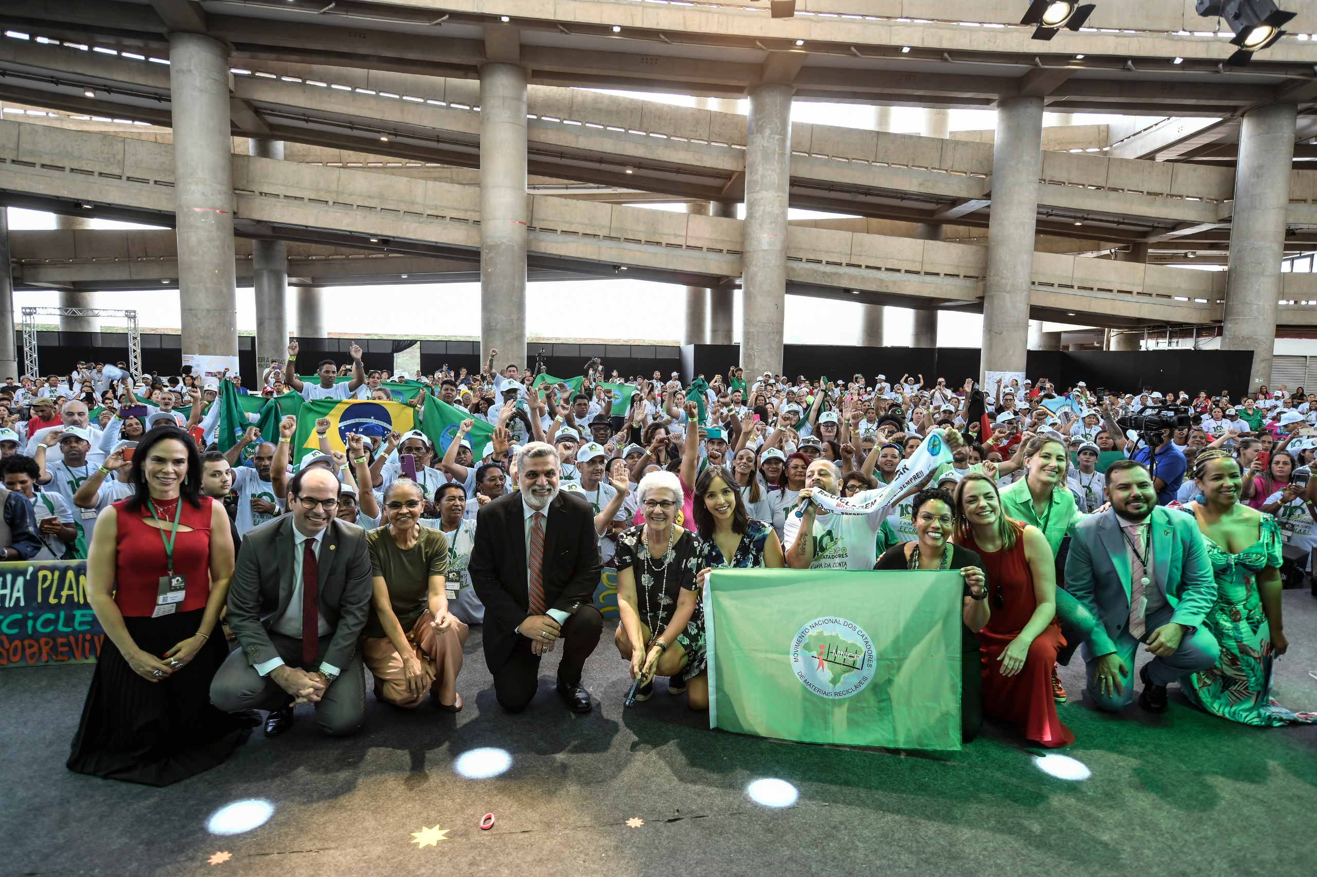 Várias pessoas agachadas, incluindo as autoridades e a categoria de catadores. Ao fundo uma parte da estrutura do estádio.